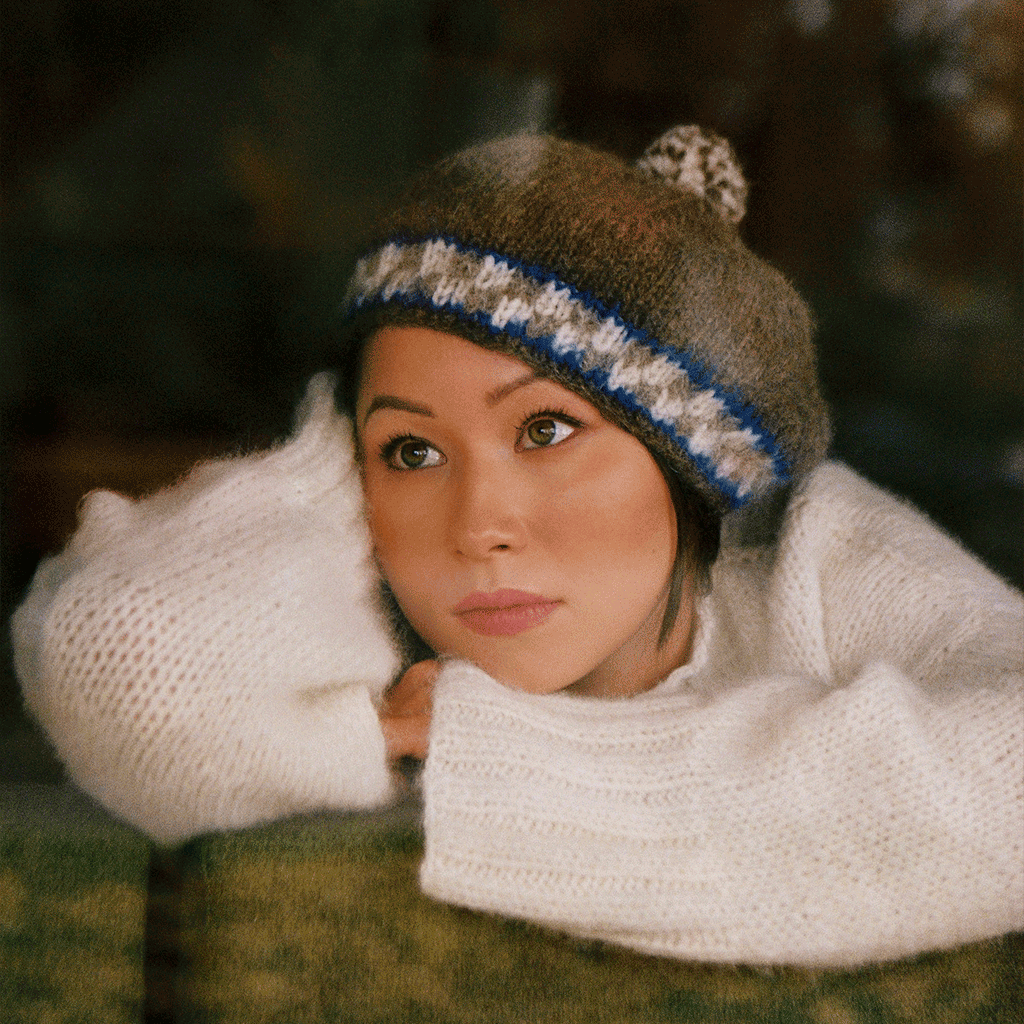 Woman wearing a grey, blue and white beret hat with a white sweater laying on a couch looking up