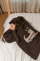 Buster Brown Best Buds hand-knit sweater worn lying on a bed, showing the cream dog and bird graphic on the back
