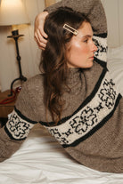 Woman lounging on a white bed in the hand knit gray flower sweater, showing the Fair Isle style flower band