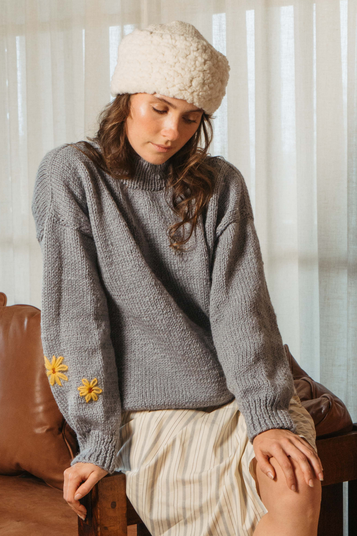 Banjo Blue Best Buds hand-knit sweater worn with a cream knit hat, seated on a leather bench, embroidered flowers on the sleeve